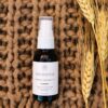 En lille flaske Innersense Harmonic Healing & Treatment Oil Skin, Scalp & Hair 25ml sidder på en tekstureret brun overflade, ledsaget af hvedestilke.