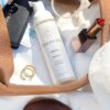 Forskellige ting, herunder en flaske Innersense Refresh Dry Shampoo - Skum til pulver 70 ml, læbestift, solbriller, ringe, et pas og andre vigtige rejseartikler er arrangeret på en overflade.