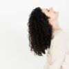En person med krøllet mørkt hår vipper hovedet tilbage og ser opad, iført en lys langærmet skjorte. Deres lokker fremstår frisk rensede, måske takket være Innersense Color Awakening Hairbath 295ml (sulfatfri), sat mod en almindelig hvid baggrund.