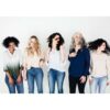 Fem kvinder med forskellige hårteksturer og -typer står på rad og række mod en hvid baggrund og bevæger legende deres hoveder og hår. De bærer afslappede toppe og jeans, der viser den ubesværede stil opnået med Innersense I Create Finish - Shine Spray 295ml.