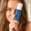 En kvinde med fregner holder en blå flaske COOLA Full Spectrum 360° Refreshing Water Mist SPF 15 50ml op i nærheden af hendes ansigt, hvilket delvist skjuler det ene øje.