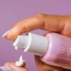 Håndpresser COOLA Dew Good Illuminating Serum Sunscreen med Probiotic SPF 30 fra en pumpeflaske på en finger mod en lilla baggrund.
