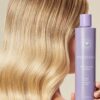 En person med bølget blond hår holder en 946 ml lilla flaske Innersense Bright Balance Hairbath, en shampoo.