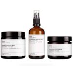 Fire hudplejeprodukter mærket "Evolve Daily Skin Essentials Trio" inklusive en rensende smelte, fugtmist, ansigtscreme og omega-serum, arrangeret på en hvid baggrund.