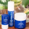 Tre COOLA hudplejeprodukter med blå beholdere: COOLA Refreshing Water Stick SPF 50 - 22 g, Refreshing Water Mist og Refreshing Water Cream, vises på et bord ved siden af en klar glaskande med citronskiver.