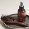 En John Masters Organics Combo Paddle Brush - Mini og en flaske John Masters Organics leave-in conditioning mist placeres på en rund, grå bakke.